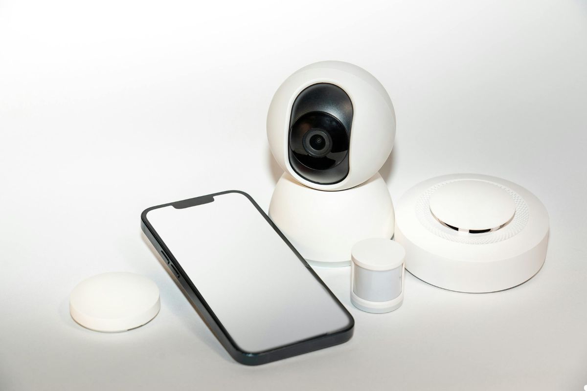 Smart home security camera, smartphone, and smoke alarm arranged on a white background, showcasing home safety technology.