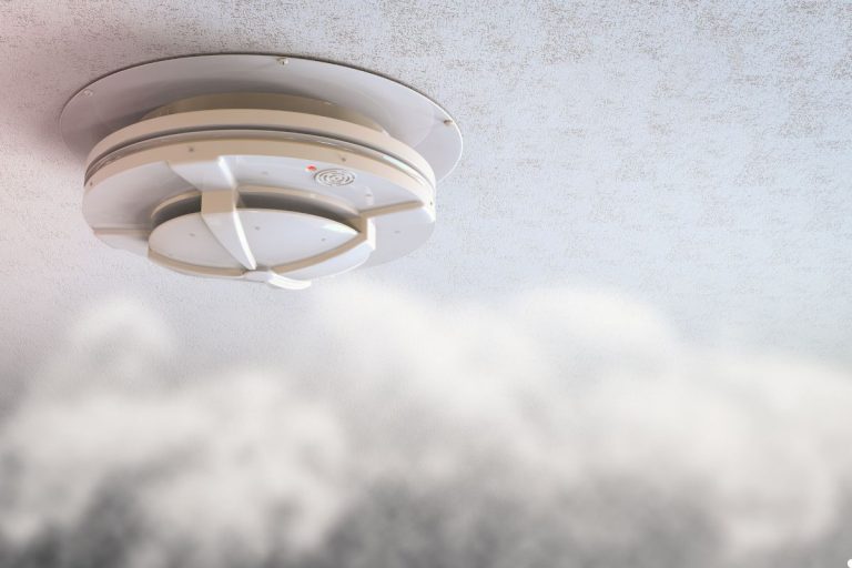 A smoke detector mounted on a ceiling with a soft cloud visible in the background.
