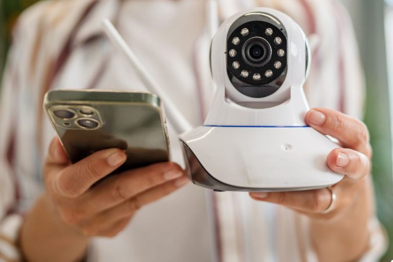 A woman stands holding a smartphone, with a security camera visible in the background, showcasing modern technology use.